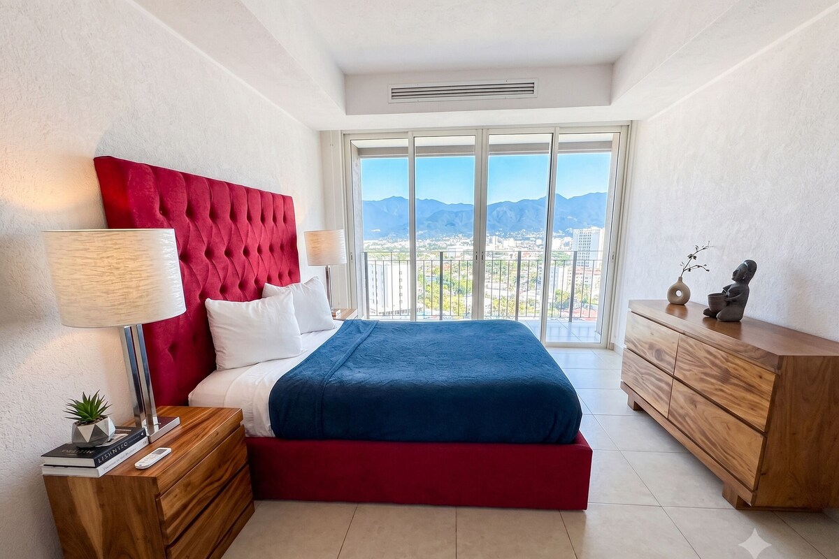 A bedroom with a bed, a nightstand, a dresser, a lamp, and a view of the city and mountains.