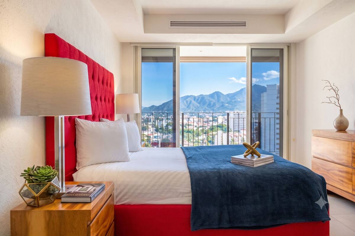 Una habitación de hotel con cama, mesita de noche, lámpara, planta y vistas a las montañas.