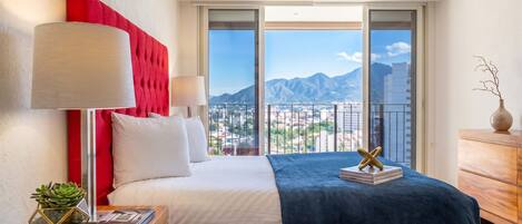 Una habitación de hotel con cama, mesita de noche, lámpara, planta y vistas a las montañas.