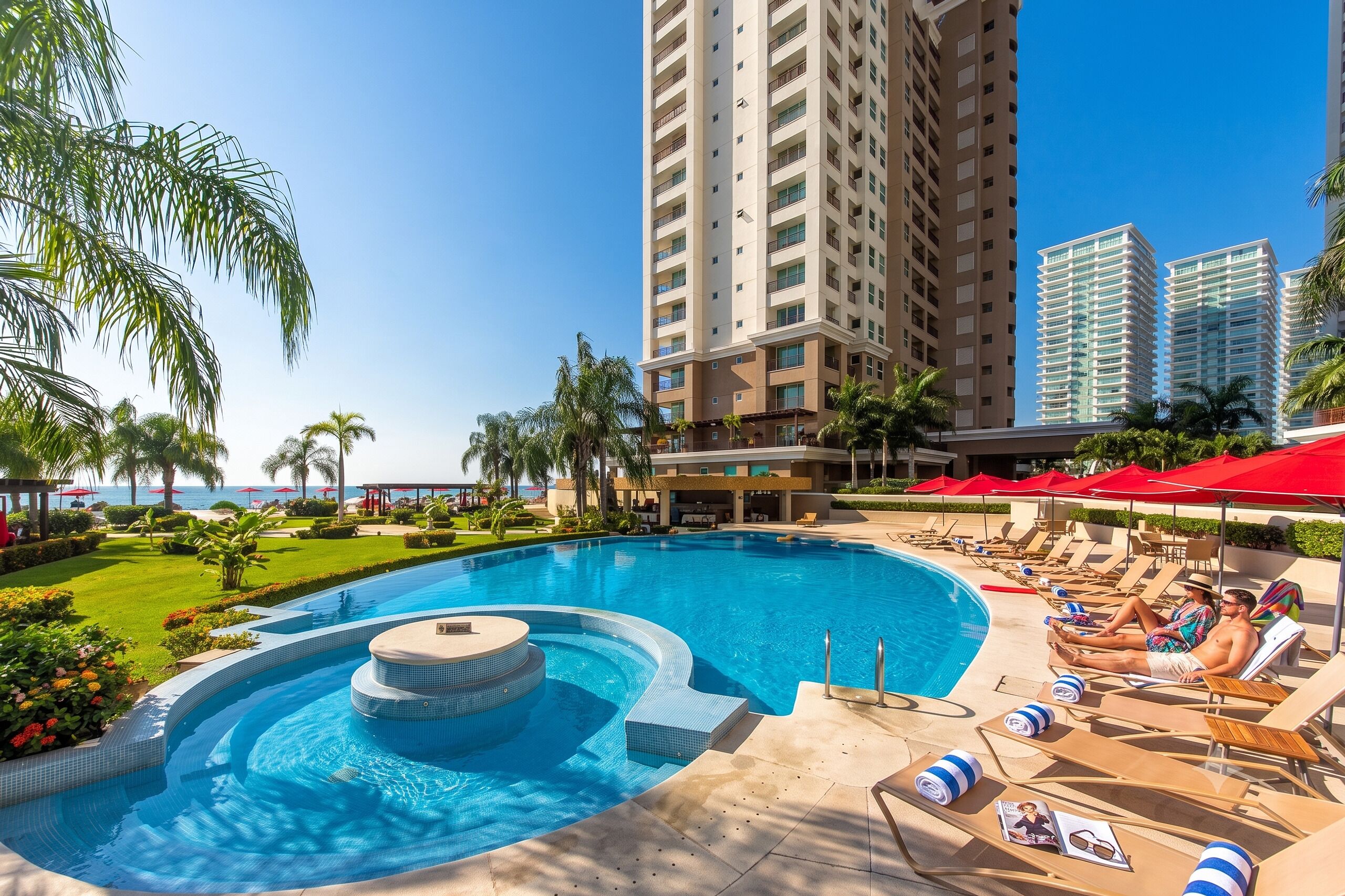 Una piscina con sillas de descanso y sombrillas, rodeada de palmeras y vista a edificios altos.