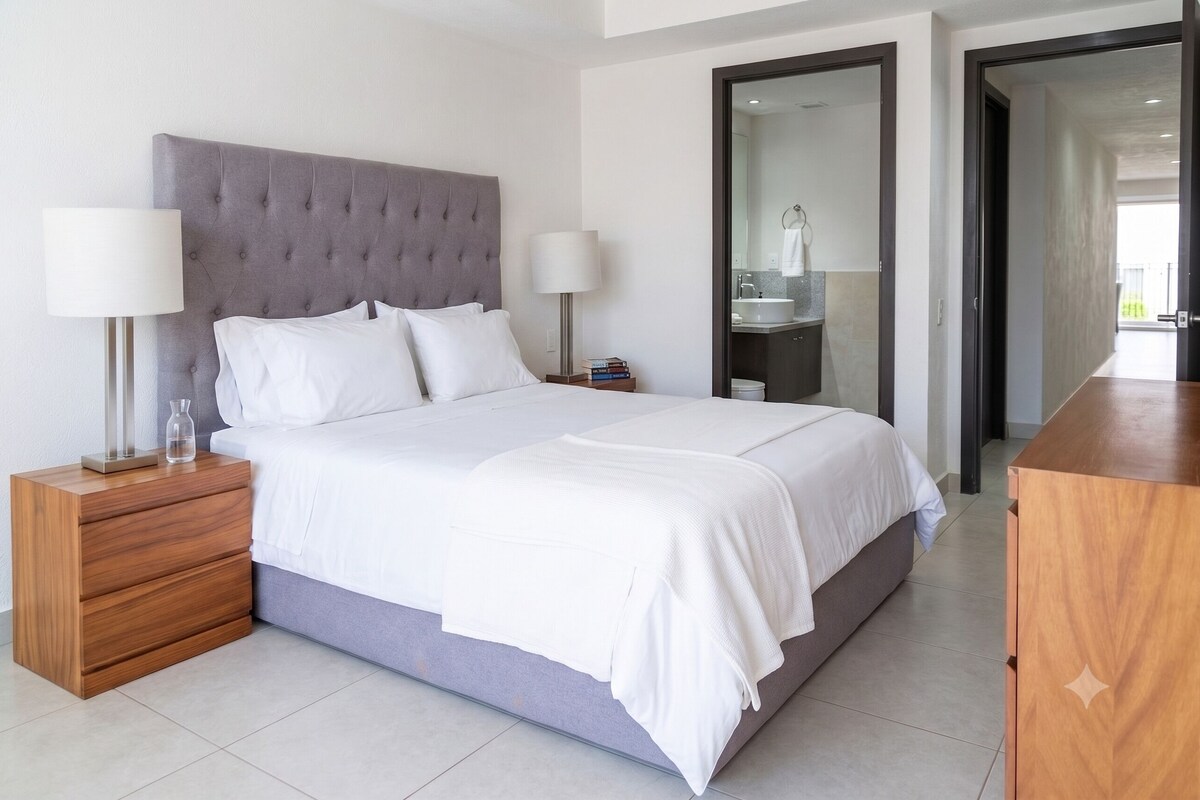 A modern hotel room with a large bed, two bedside tables with lamps, a wooden nightstand, and a view of the bathroom and hallway.