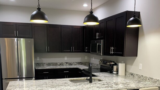 Una cocina moderna con gabinetes oscuros, electrodomésticos de acero inoxidable, una encimera de granito y tres taburetes de bar.