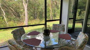 Una mesa redonda de vidrio con cuatro sillas, cada una con un cojín estampado, en una habitación con vista a un espacio verde y árboles.