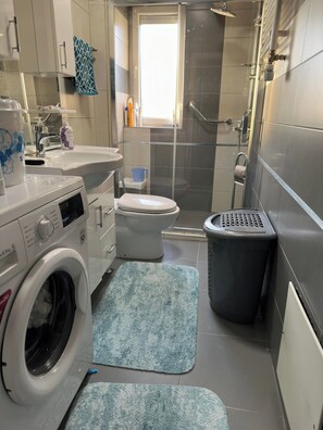 A compact bathroom with a washing machine, toilet, shower, and a trash bin. - Gorgeous 1-bedroom apartment with AC in lively Prishtinë (Prishtinë)