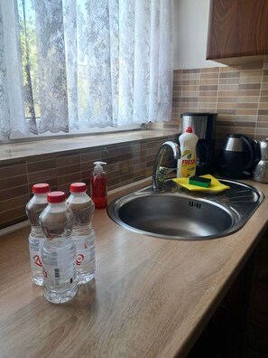 Eine Küche mit einer Spüle aus Edelstahl, einem Fenster mit Spitzenvorhängen und verschiedenen Küchengegenständen, darunter Flaschen und eine Kaffeemaschine.