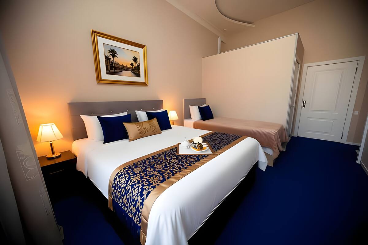 Habitación de hotel con una cama grande, otra más pequeña, una mesita de noche con lámpara, un cuadro en la pared y una puerta.