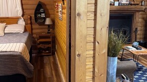 A cozy wooden cabin room with a bed, a small table, a fireplace, and a shelf with various items. - The Willows on Table Rock Lake, 1 Bed / 1 Bath Log Cabin, Pool, Hot Tub & Dock (Shell Knob)