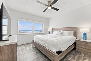 A bedroom with a bed, a ceiling fan, a window with a view of the ocean, and a desk with a lamp.