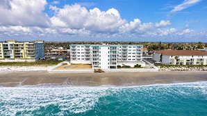 Un hotel frente a la playa con varios pisos, una playa arenosa en primer plano y vista al océano.