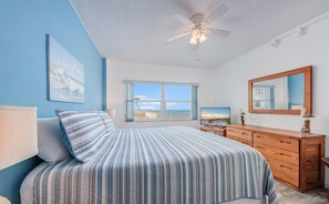 A bedroom with a bed, a TV, a dresser, a ceiling fan, and a large window with a view of the beach.