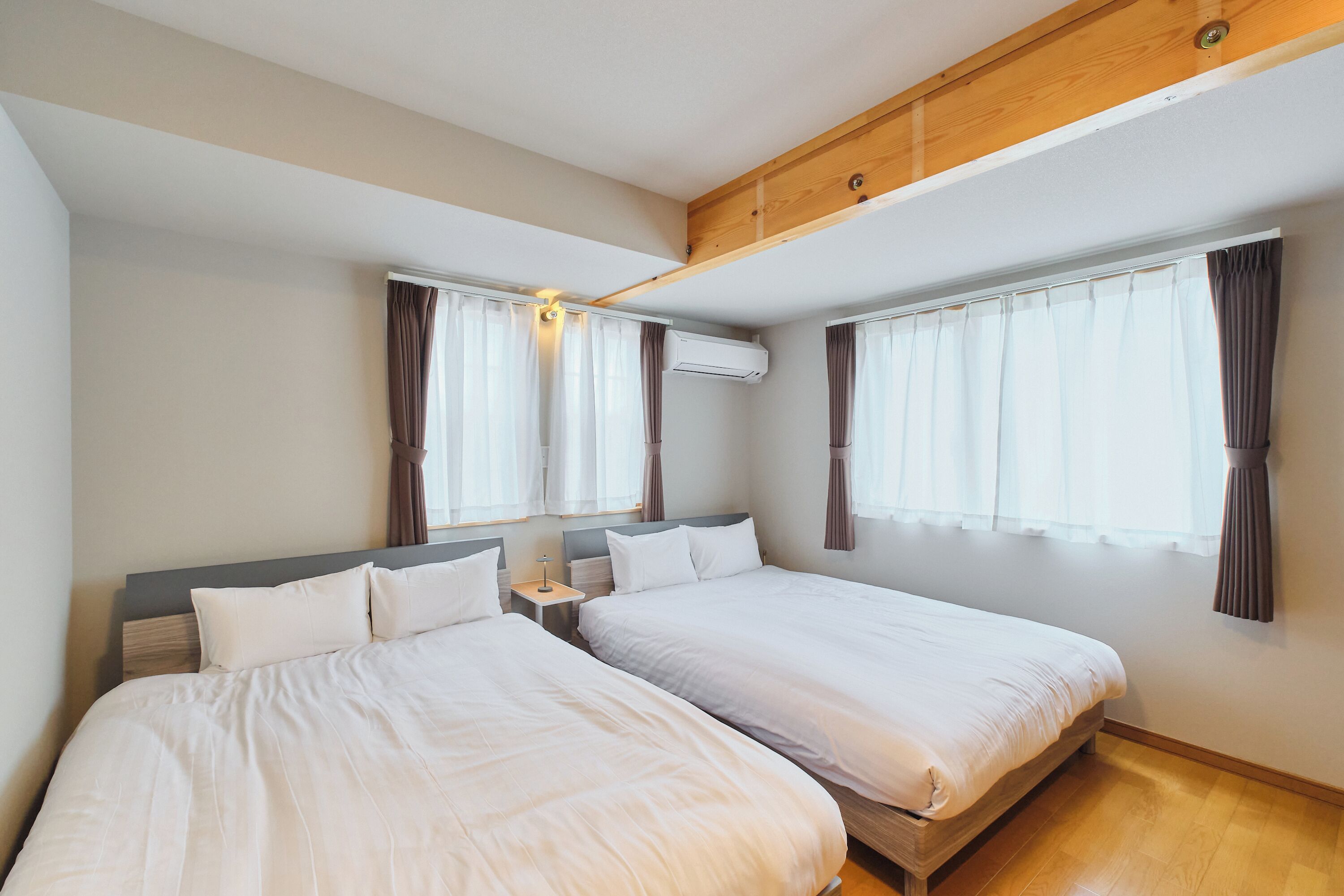 Habitación de hotel con dos camas, una viga de techo de madera, un equipo de aire acondicionado y cortinas en las ventanas.