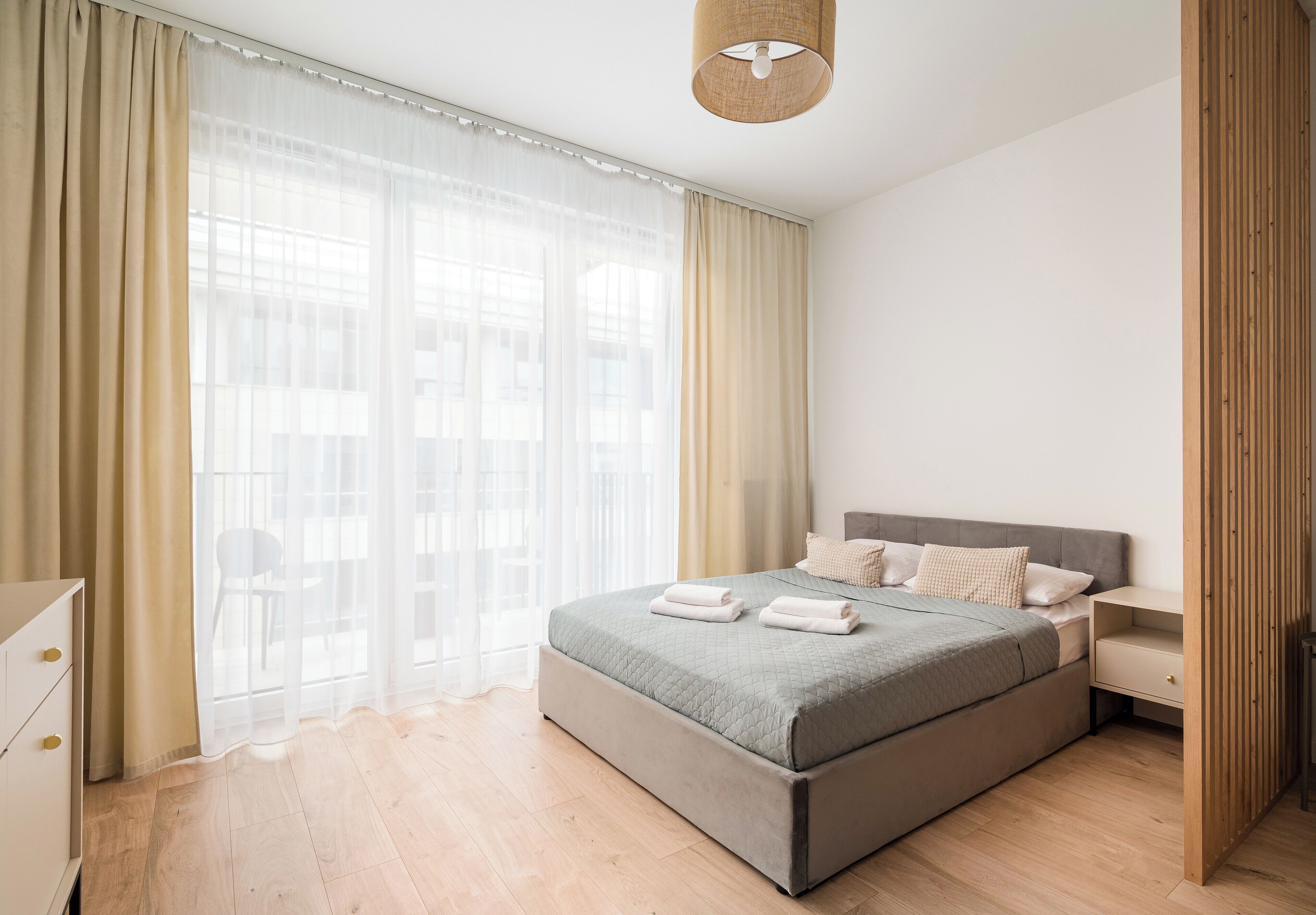 Un dormitorio moderno con una cama, cortinas, una mesita de noche y una lámpara.