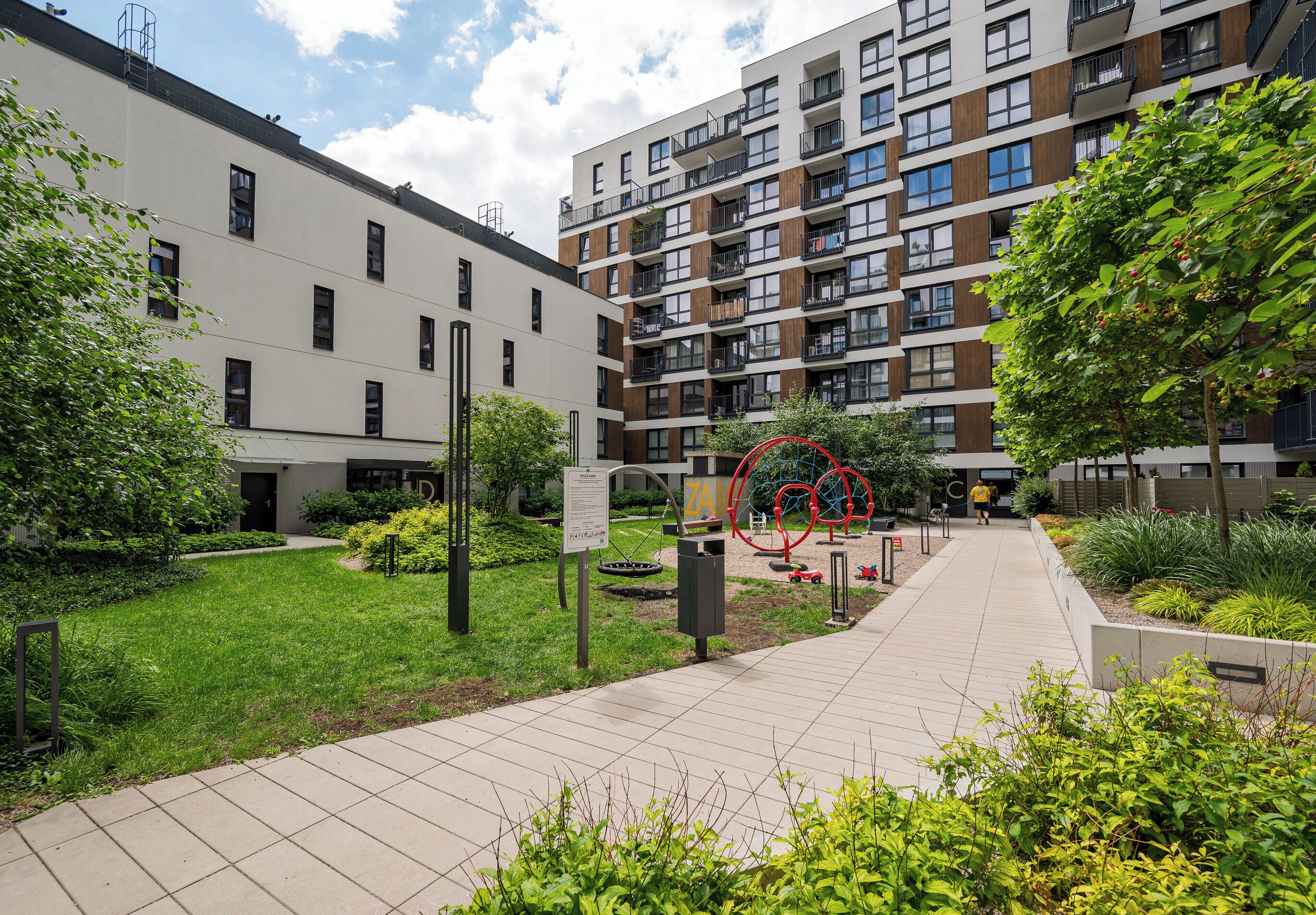 Un immeuble d’appartements moderne doté de nombreux balcons, d’un jardin aménagé et d’une sculpture circulaire rouge.