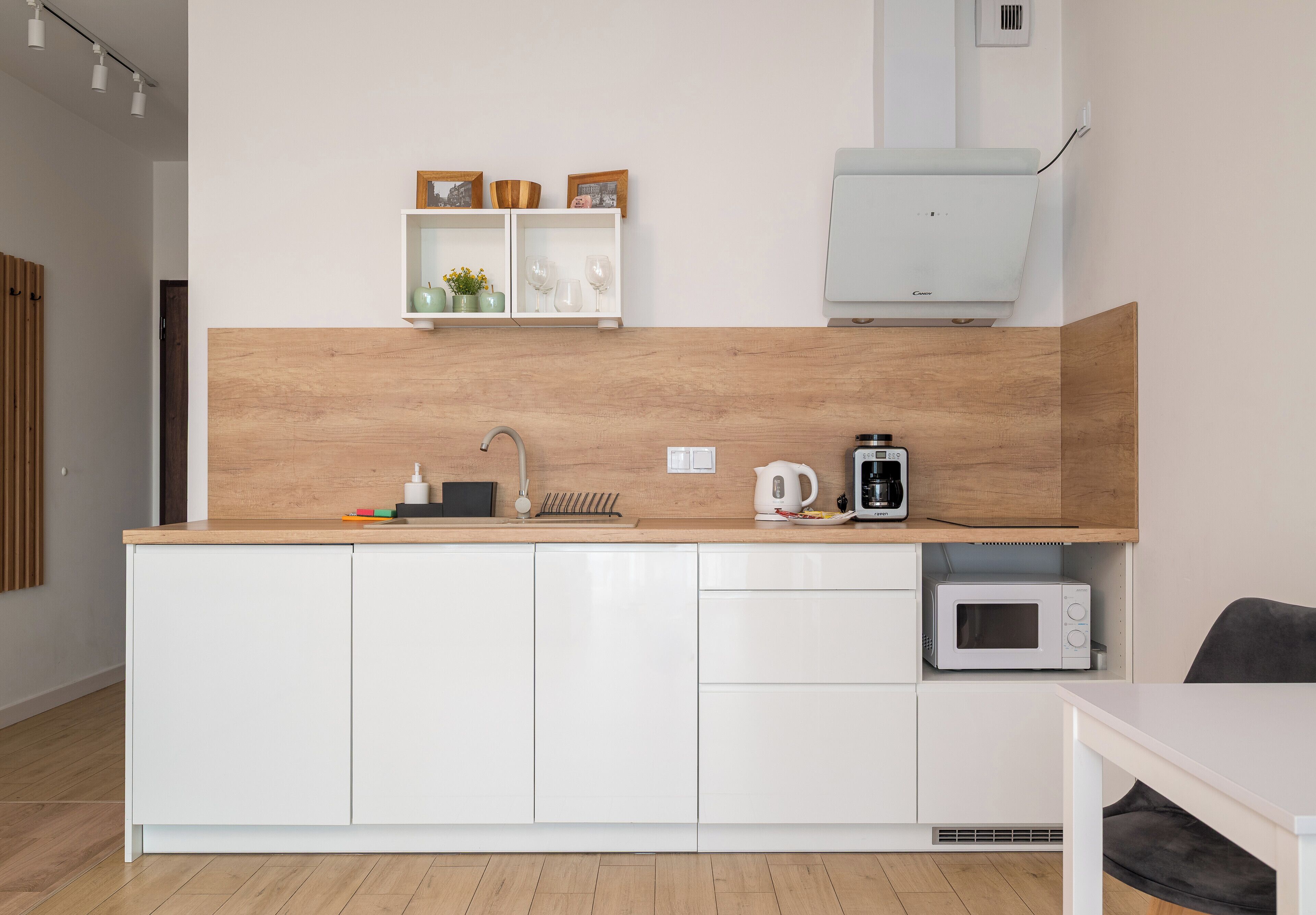Une cuisine moderne avec des meubles blancs, un plan de travail en bois, un four à micro-ondes, une machine à café et une étagère ornée d’objets décoratifs.