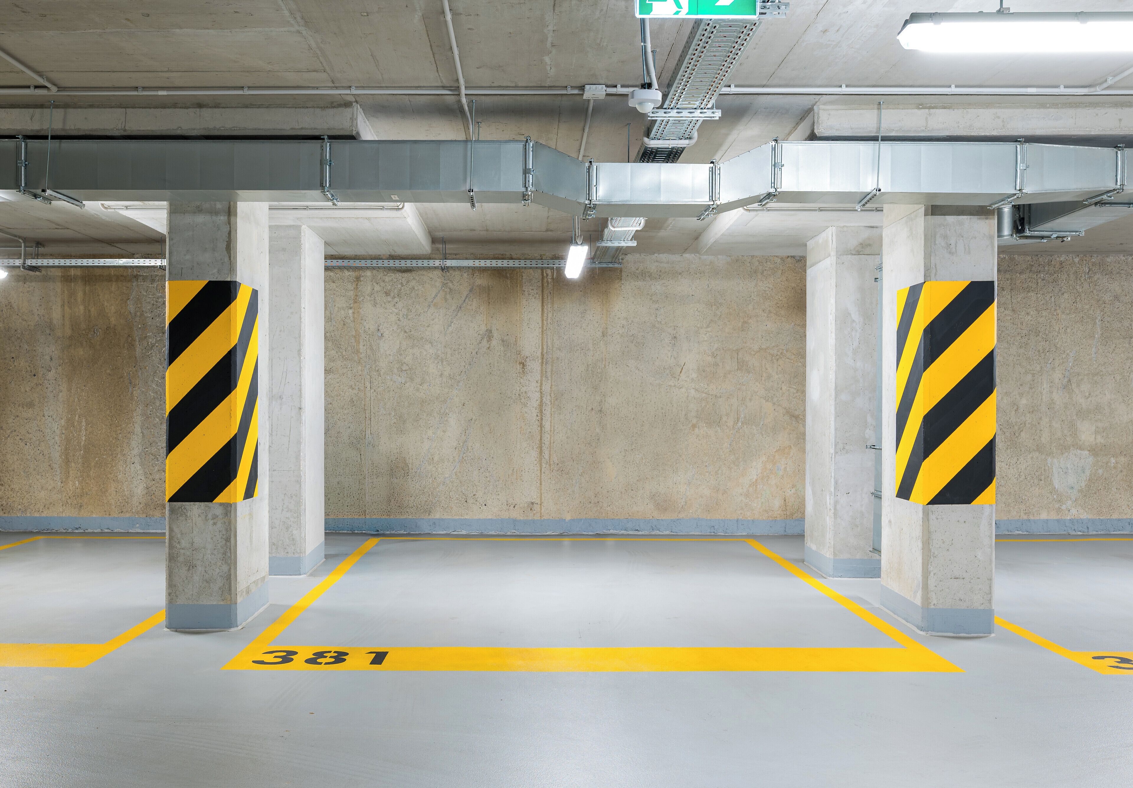 Een parkeergarage binnen met betonnen pilaren, gele parkeerlijnen en een nooduitgangbord.