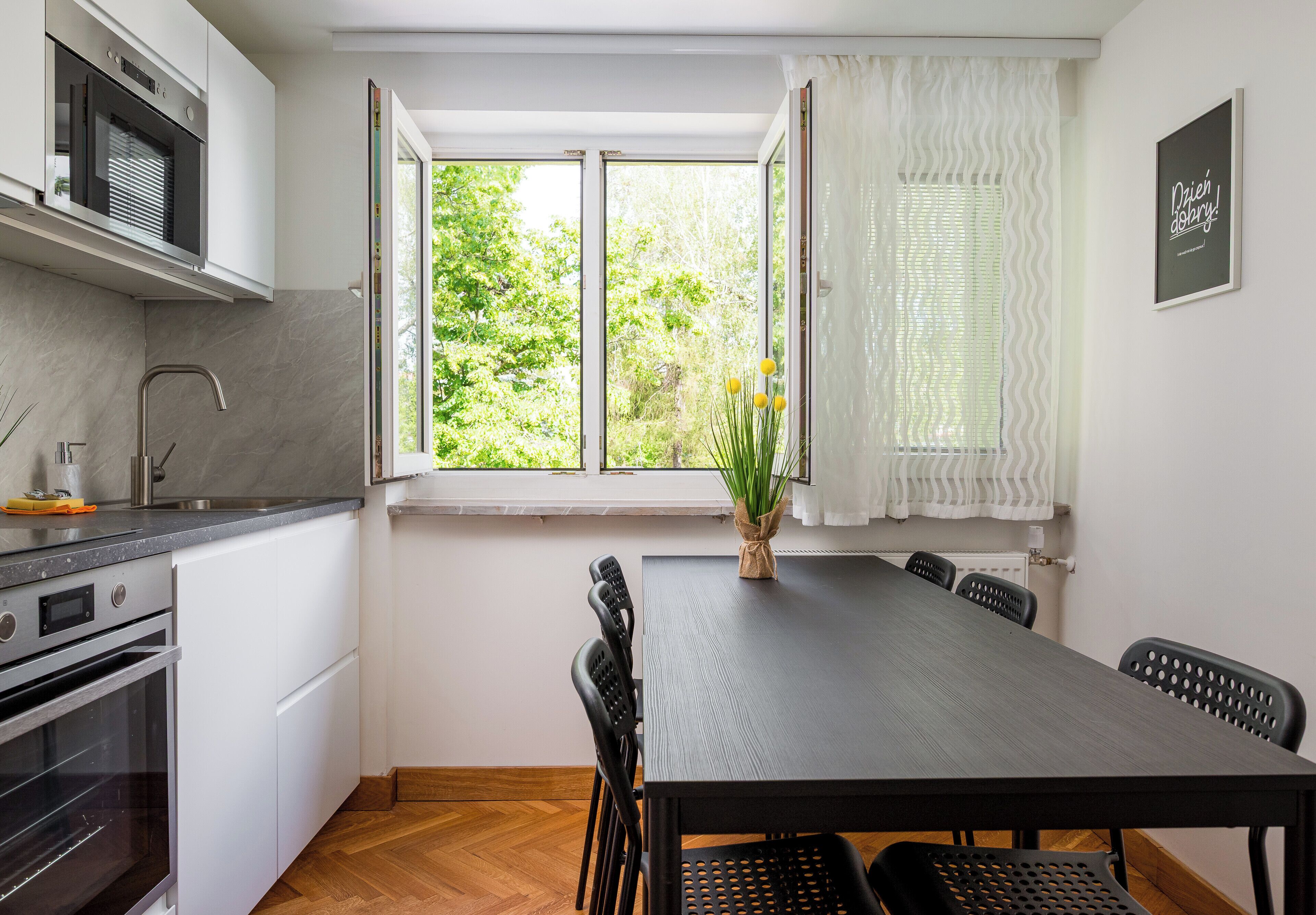 A modern kitchen with a dining area, a large window, and a view of greenery.