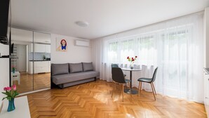 A modern living room with a sofa, dining table, chairs, a vase with flowers, and a large window with sheer curtains. - Metro Służew Apartment #6 (Warszawa)