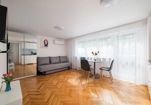 A modern living room with a sofa, dining table, chairs, a vase with flowers, and a large window with sheer curtains. - Metro Służew Apartment #6 (Warszawa)