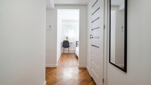A hallway with a wooden floor, white walls, a black chair, and a bed in the background. - Metro Służew Apartment #6 (Warszawa)