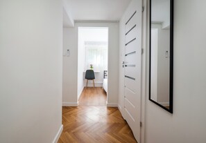 A hallway with a wooden floor, white walls, a black chair, and a bed in the background. - Metro Służew Apartment #6 (Warszawa)
