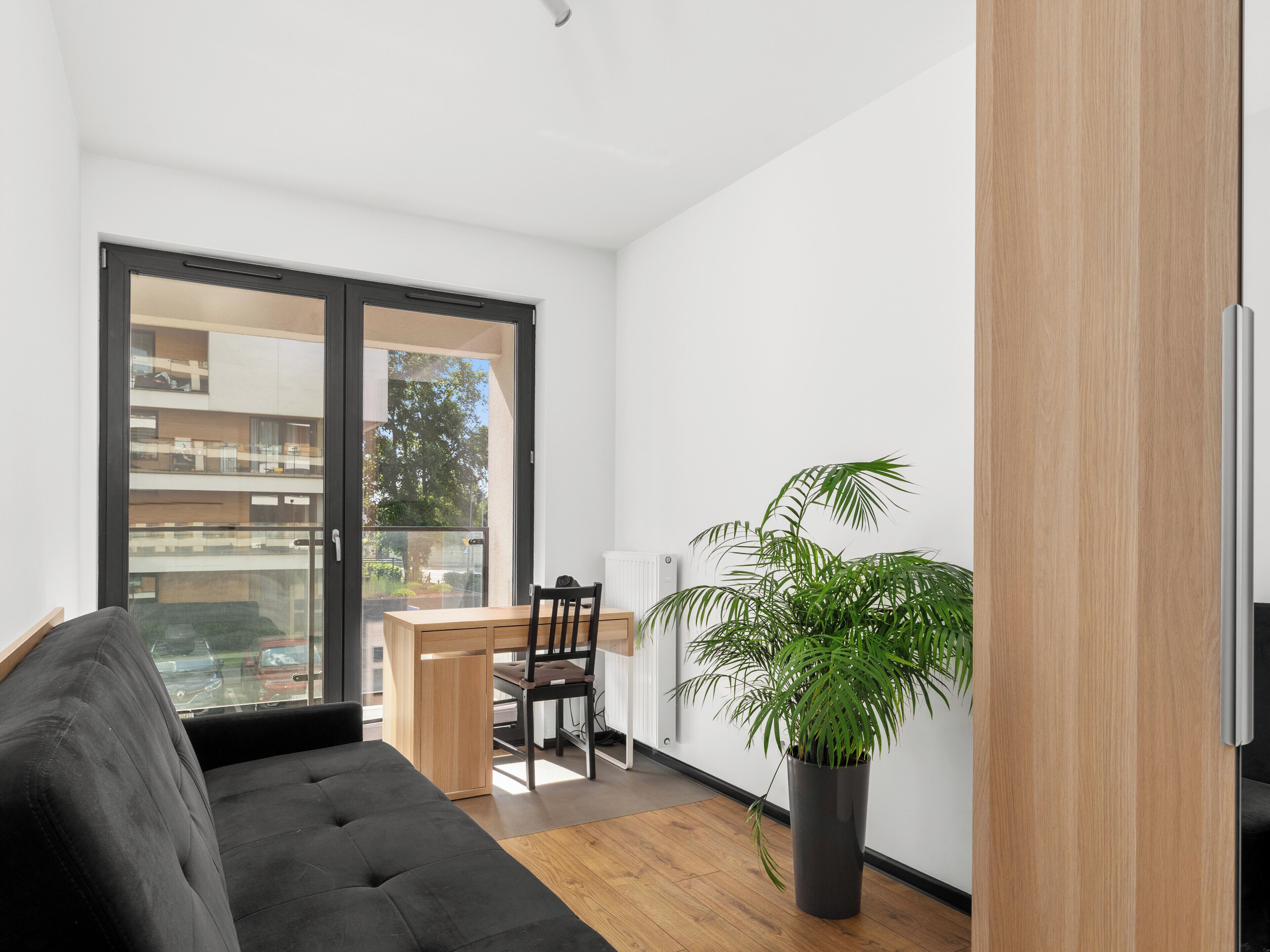 A modern apartment with a black sofa, a dining table with chairs, a potted plant, and a view through a glass door.
