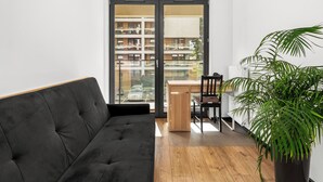 A modern living room with a black sofa, wooden flooring, a dining table with chairs, a potted plant, and a view of a multi-story building through a glass door. - Tauron Arena Harmony Apartment (Kraków)