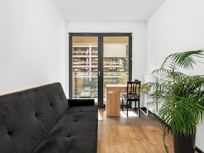 A modern living room with a black sofa, wooden flooring, a dining table with chairs, a potted plant, and a view of a multi-story building through a glass door. - Tauron Arena Harmony Apartment (Kraków)