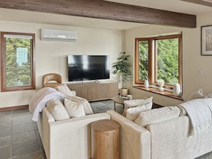 Ein Wohnzimmer mit einer Couch, einem Fernseher und Fenstern mit Blick auf Grünflächen.