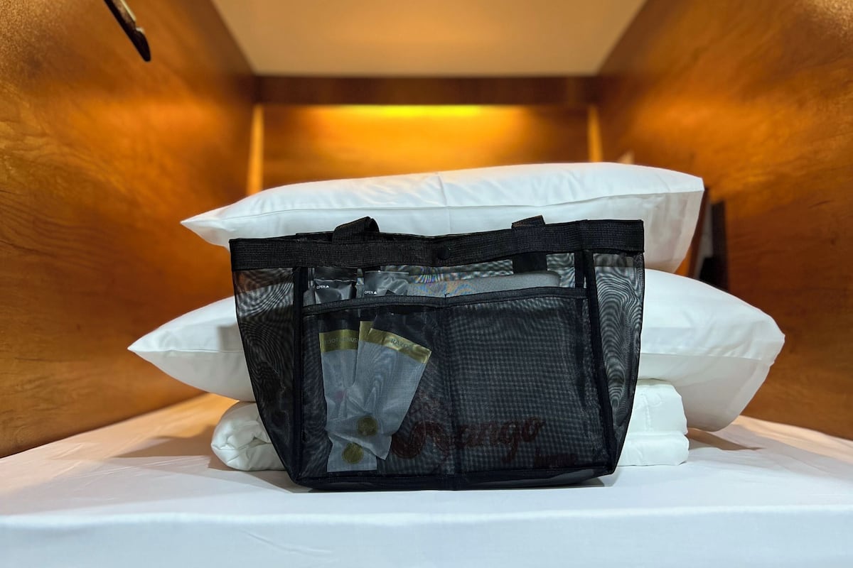 A hotel room with a bed, white pillows, a black mesh bag on the bed, and wooden walls.