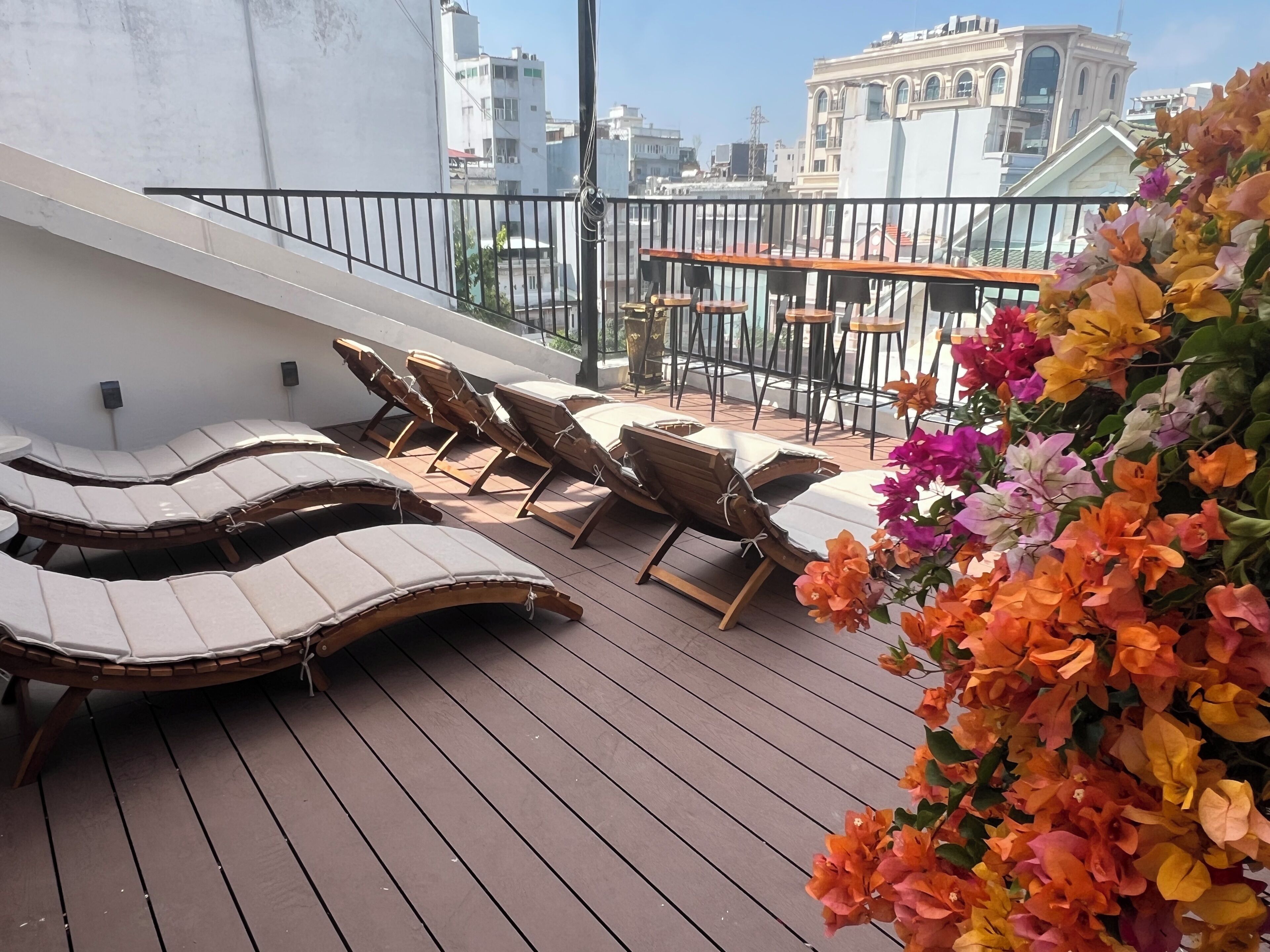 A rooftop terrace with sun loungers and a table, surrounded by a railing and overlooking a cityscape.