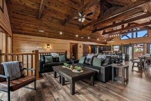 A spacious living area with a fireplace, leather sofas, wooden flooring, and a kitchen in the background. - Sleeps 30 | Heated Pool | Arcade, Game Room & Theater | Mtn Views (seveirville)