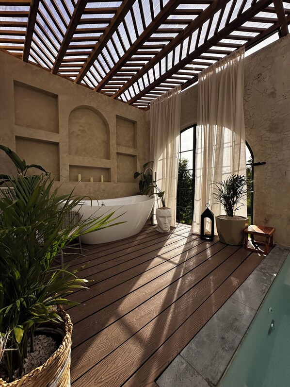 Eine Holzterrasse mit einer großen Badewanne, Topfpflanzen und Blick durch ein Rundbogenfenster.