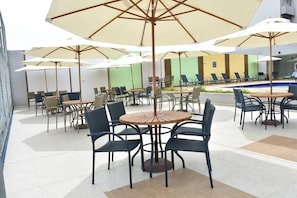 An outdoor seating area with round tables and chairs, umbrellas, and a pool in the background. - Caldas Novas - Recanto do Bosque Flat (Caldas Novas)