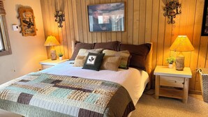 Een slaapkamer met een houten lambrisering, een bed met veel kussens, twee bedlampjes en een lijst met een afbeelding erboven.
