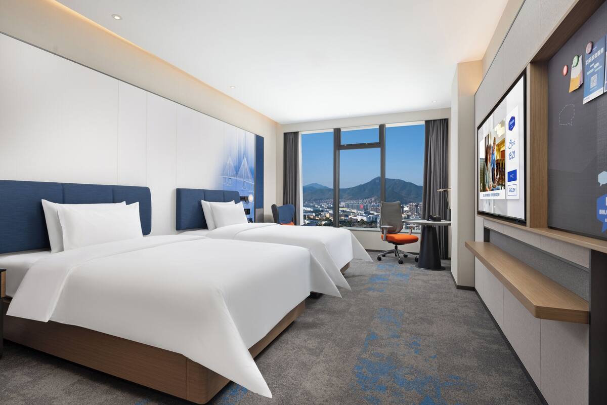 Habitación de hotel con dos camas, un escritorio, una silla y un ventanal con vista a la ciudad.