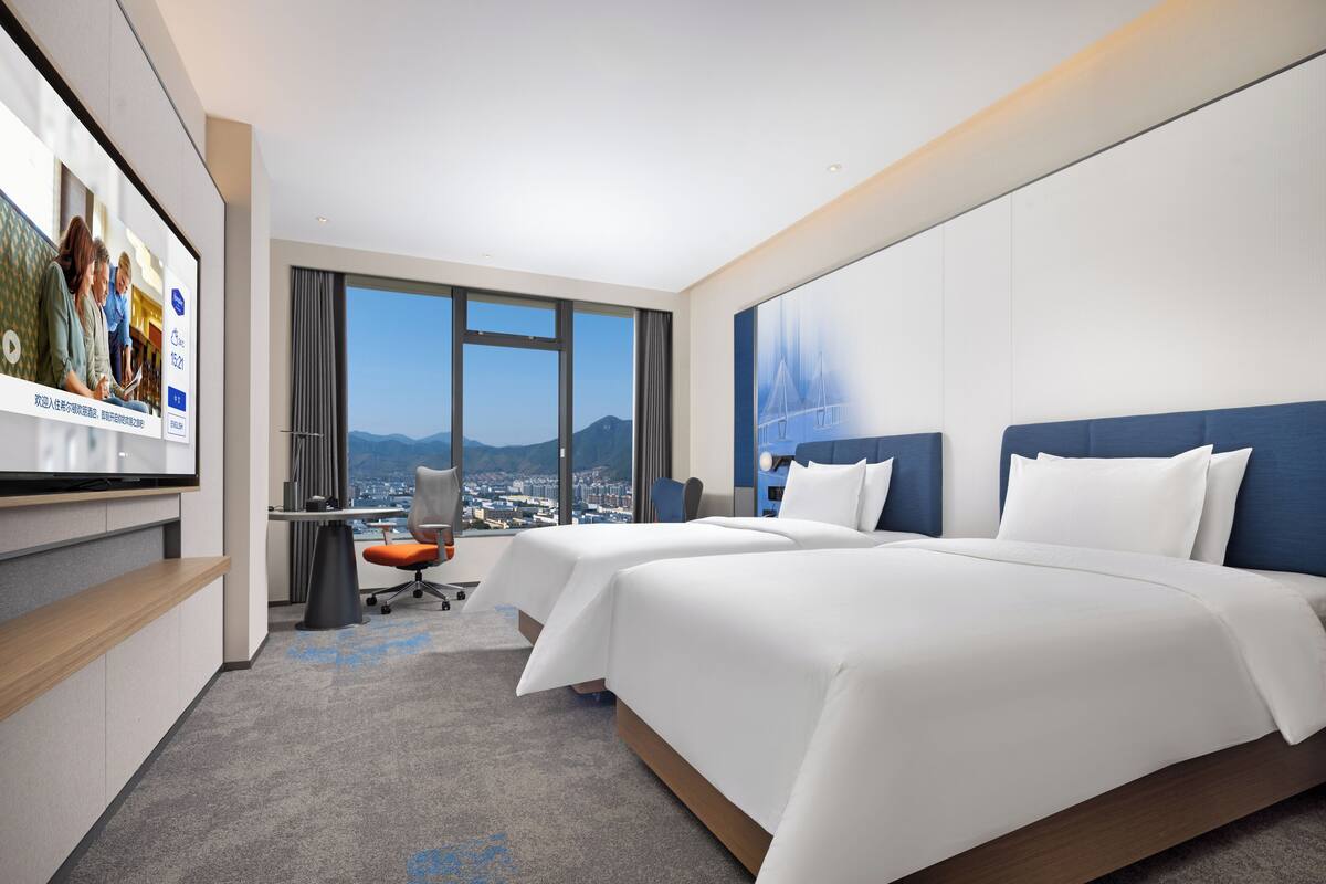 Habitación de hotel con dos camas, un escritorio, una silla, un televisor y un ventanal con vistas a la ciudad.