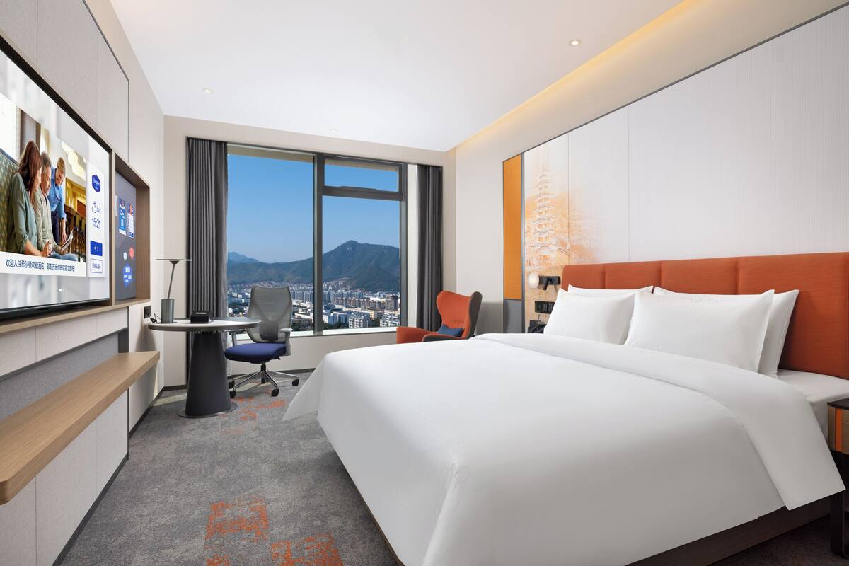 Habitación de hotel con una cama grande, un escritorio, una silla, un televisor y vistas a la ciudad y montañas.
