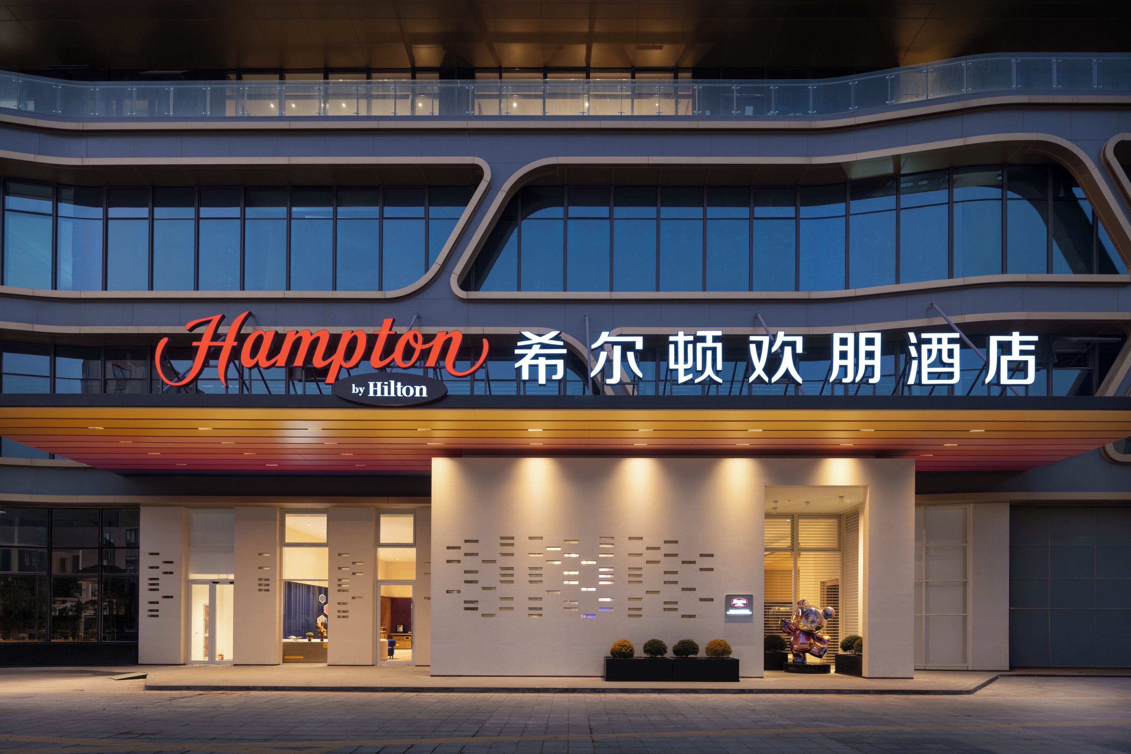 A modern hotel with a prominent 'Hampton by Hilton' sign, large windows, and a clean facade.