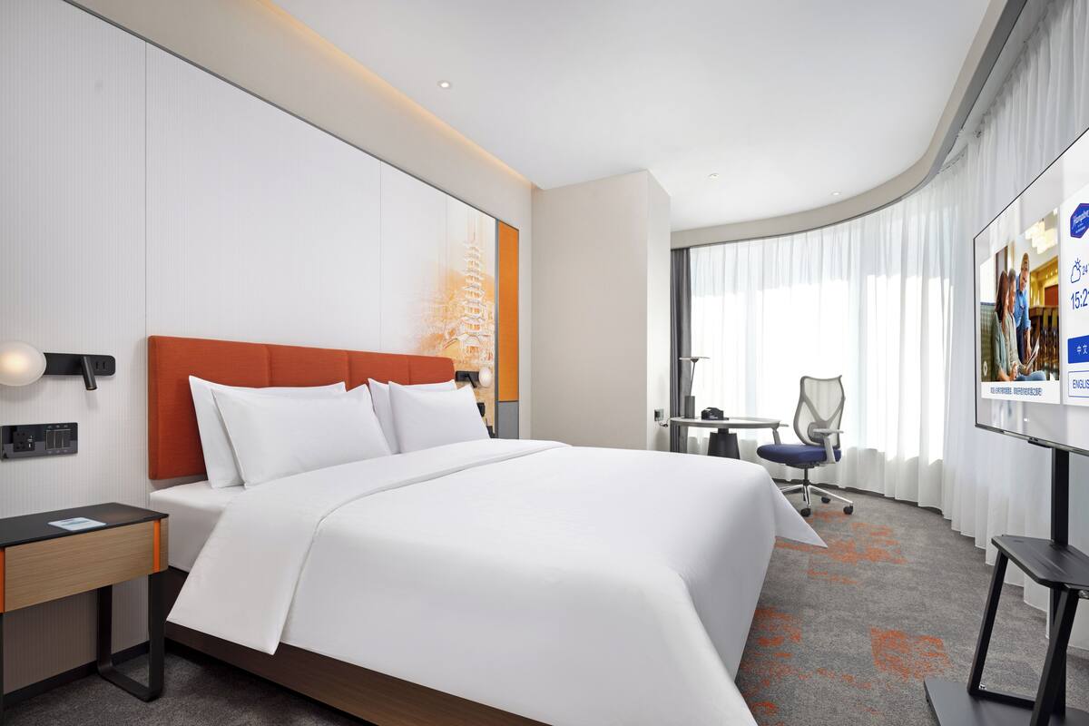 Una habitación de hotel con una cama grande, un escritorio con silla, televisión y ventana con cortinas.