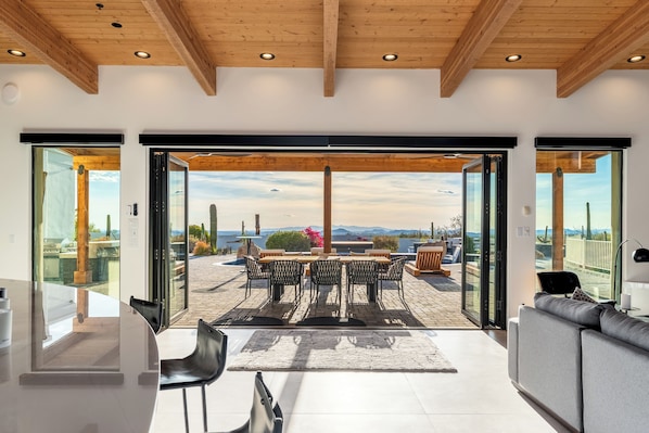 A modern interior with a dining area, a sofa, and a view through glass doors. - NEW SWSS 8BR Desert‑Modern Estate | Pool, Hot Tub, Golf Simulator, Cold Plunge, Putting Green (Scottsdale)