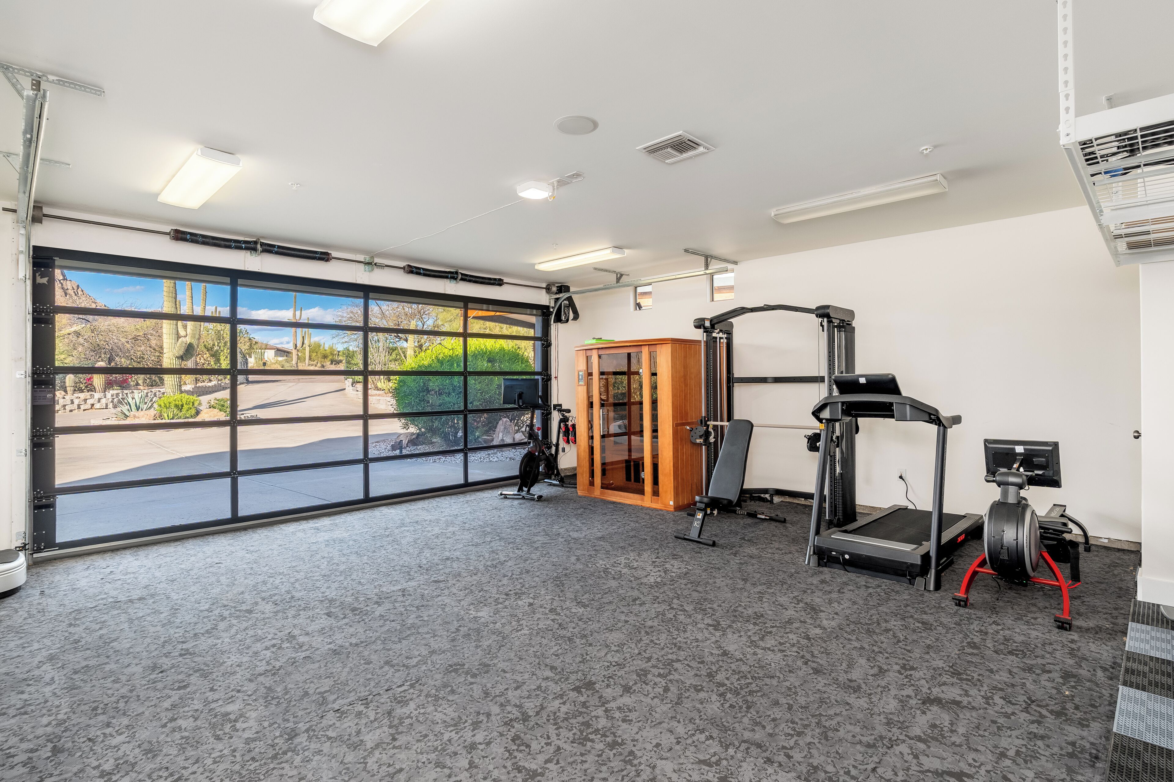 A gym with a treadmill, elliptical, and weight machine, large windows with a desert view, and a door leading outside.