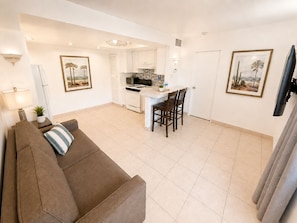 A modern living room with a brown sofa, a dining area with two chairs, a kitchen with white cabinets, and a TV mounted on the wall.