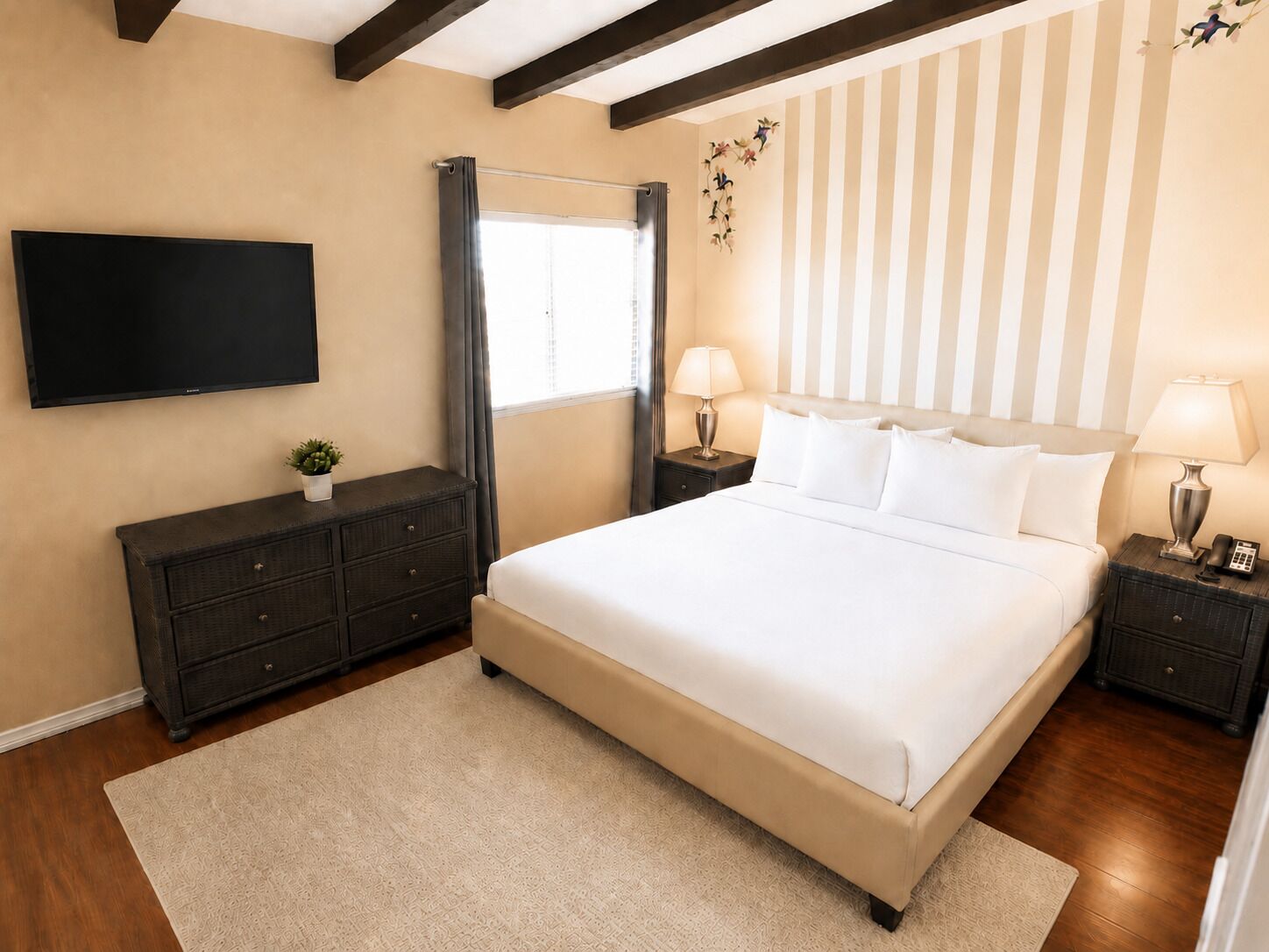 A bedroom with a large bed, a flat-screen TV, a dresser, two bedside tables with lamps, and a window with curtains.
