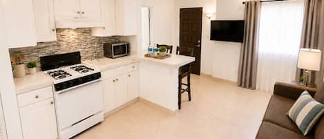 Una cocina moderna con armarios blancos, una estufa y un microondas. Hay una zona de comedor con un sofá marrón y un televisor montado en la pared.
