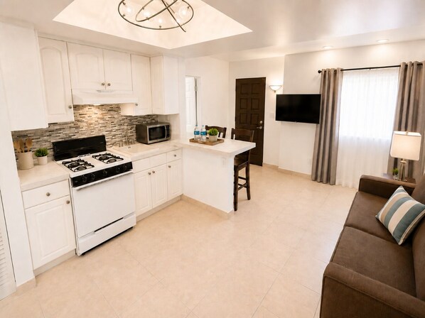 A modern kitchen with white cabinets, a stove, and a microwave. There's a dining area with a brown sofa and a TV mounted on the wall.
