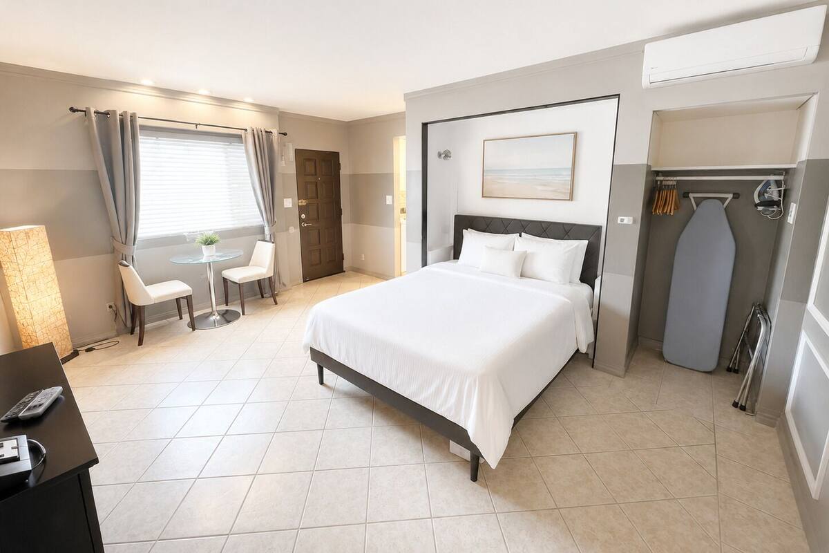 Una habitación de hotel con cama, escritorio con teléfono, silla, mesita con planta y ventana con cortinas.