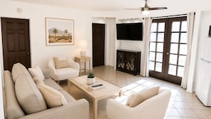 A living room with a sofa, armchairs, a coffee table, a TV, and a door. - Mykonos Desert Suites (Palm Springs)