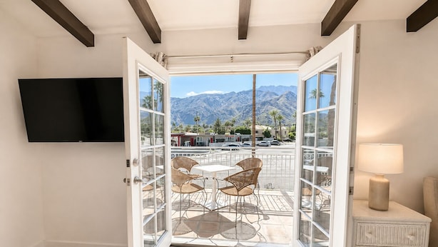 A room with a large sliding glass door leading to an outdoor patio with a mountain view, a flat-screen TV mounted on the wall, and a small white nightstand with a lamp. - Mykonos Desert Suites (Palm Springs)