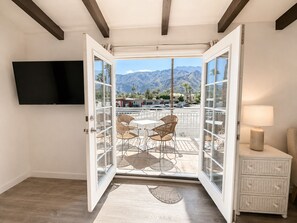A room with a large sliding glass door leading to an outdoor patio with a mountain view, a flat-screen TV mounted on the wall, and a small white nightstand with a lamp.