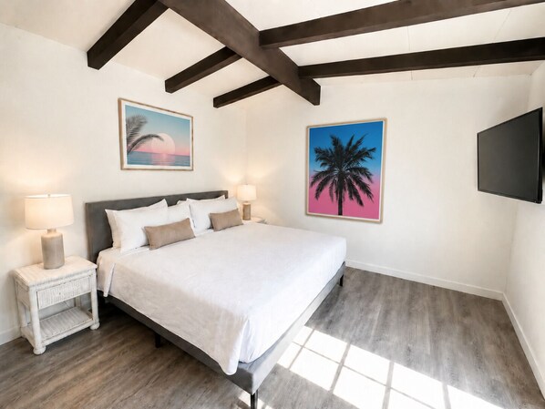 Um quarto com cama, duas mesas de cabeceira com abajures, um quadro grande de uma palmeira e uma TV fixada na parede.
